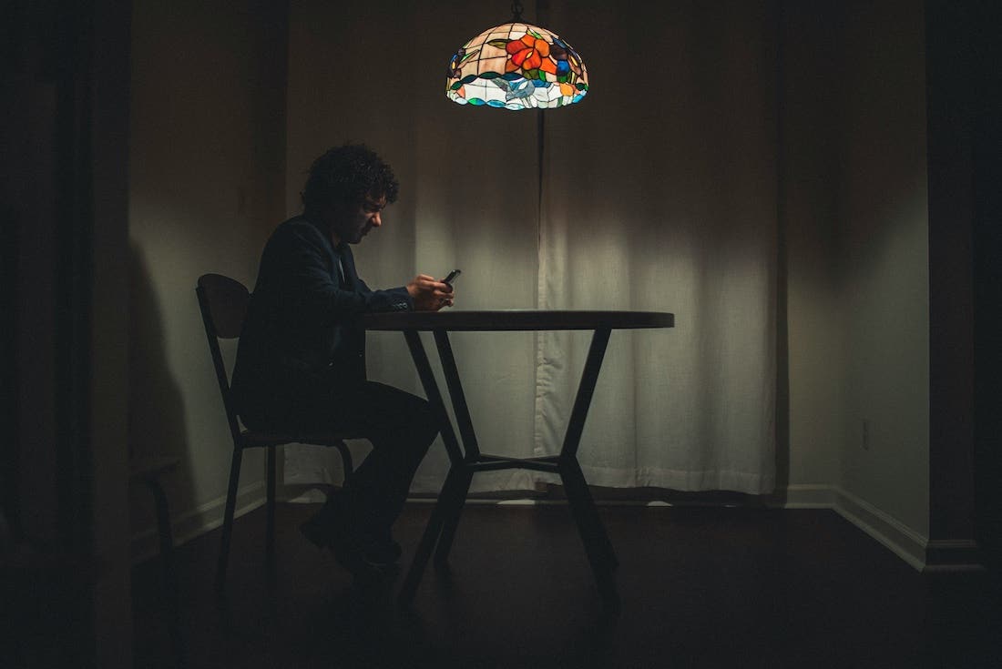 Man sitting at a table alone under a lit lamp looking at his phone